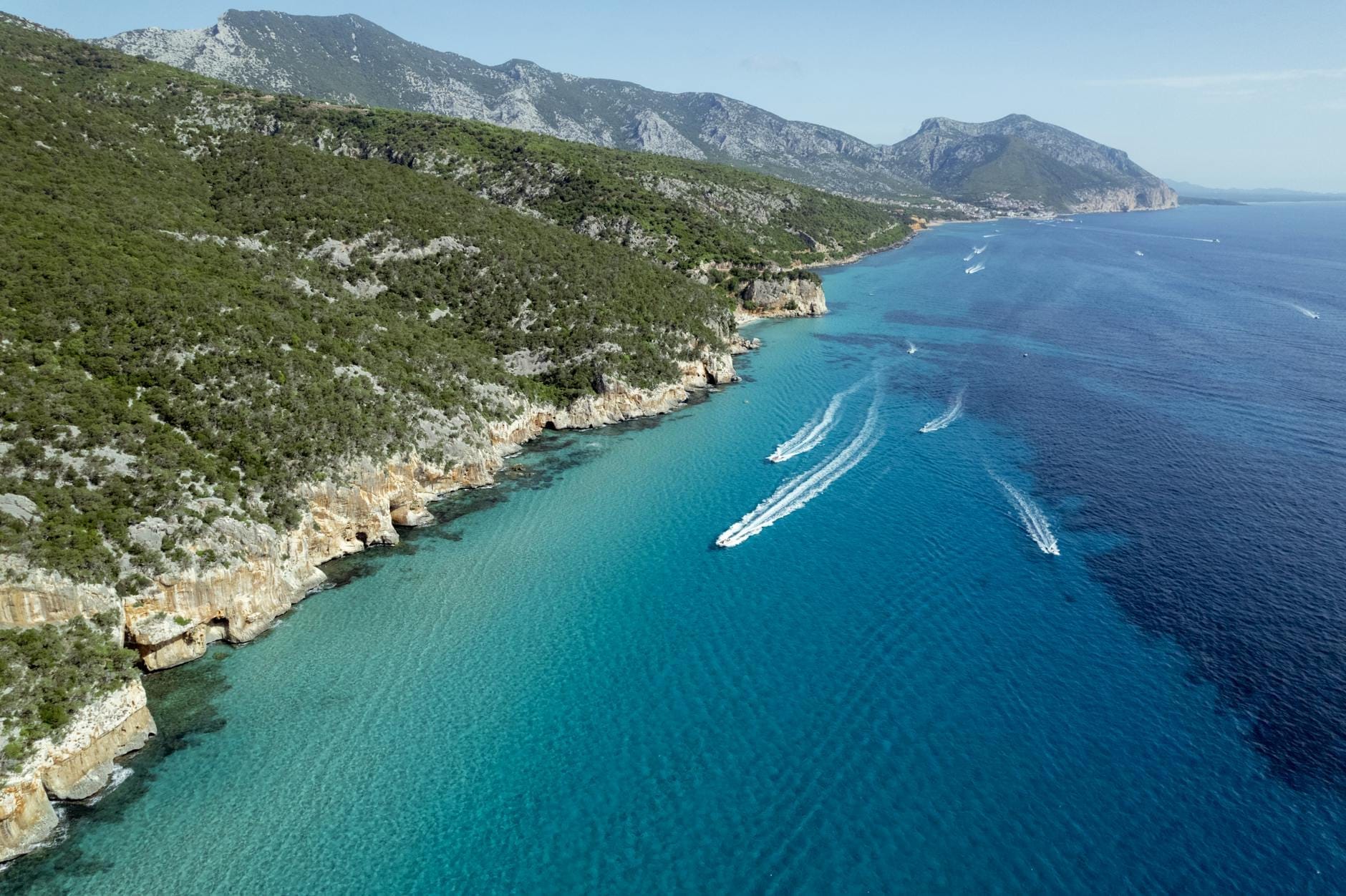 Luftaufnahme der dramatischen sardischen Küstenlinie mit türkisfarbenem Wasser, zerklüfteten Klippen und Booten, die die atemberaubenden Aussichten am Ende von Küstenwanderungen veranschaulicht.