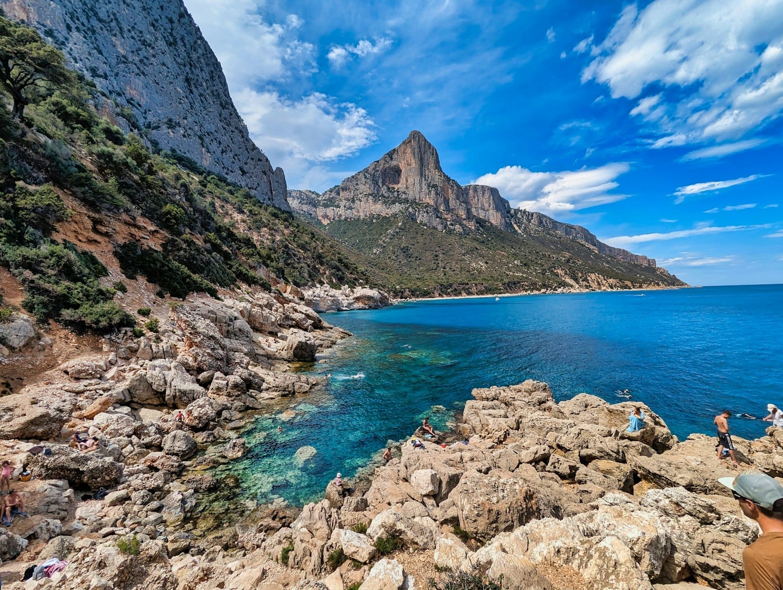 Dramatischer Kalksteingipfel hoch über türkisfarbenem Meer und felsiger Küste mit rauem Wanderpfad – Sardiniens Bergwelt und Küstenlandschaft unter blauem Himmel.