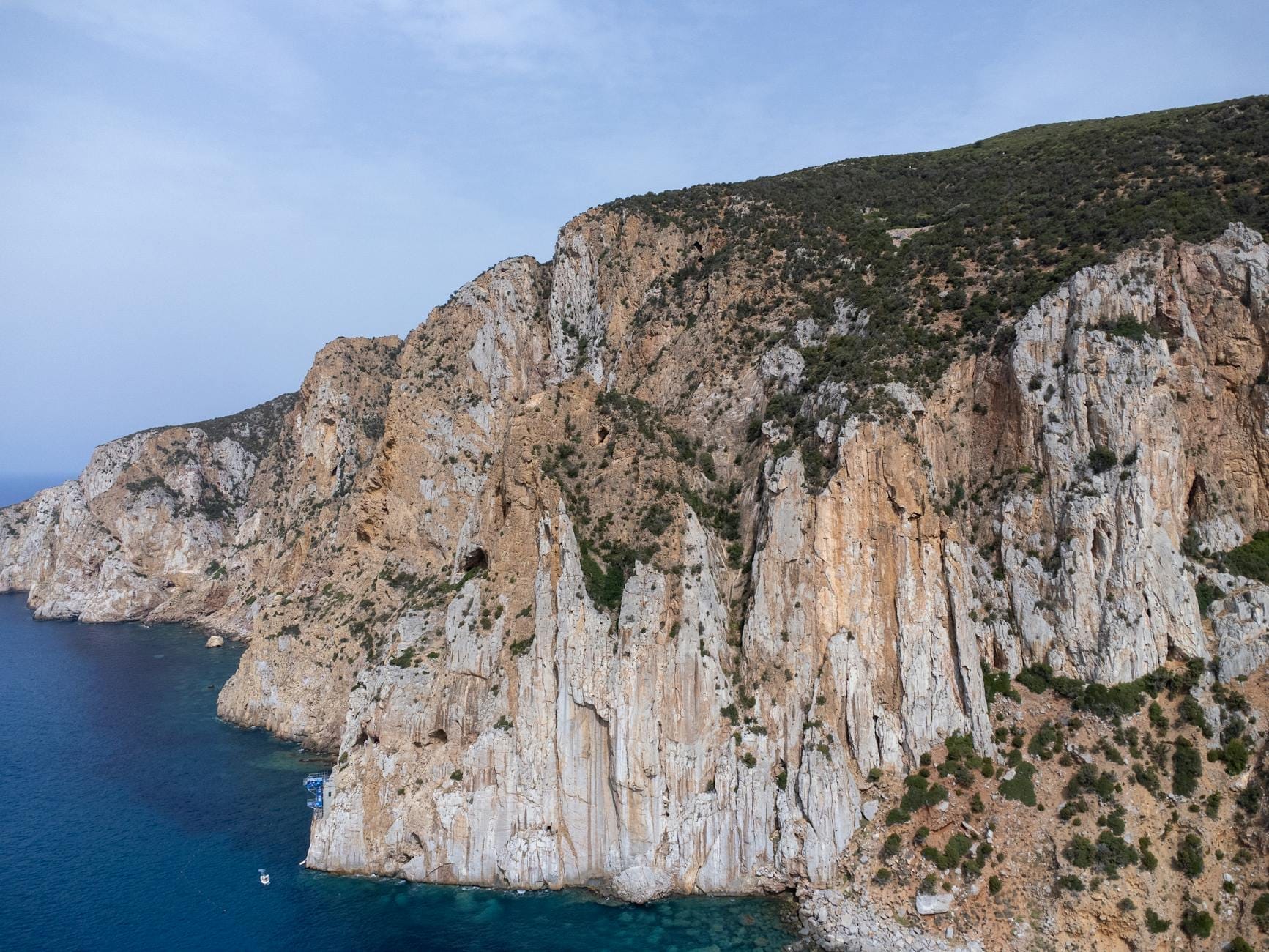 Steile, zerklüftete Klippen ragen über das tiefblaue Meer, mit spärlicher Vegetation auf abschüssigem Felsgelände unter klarem Himmel.