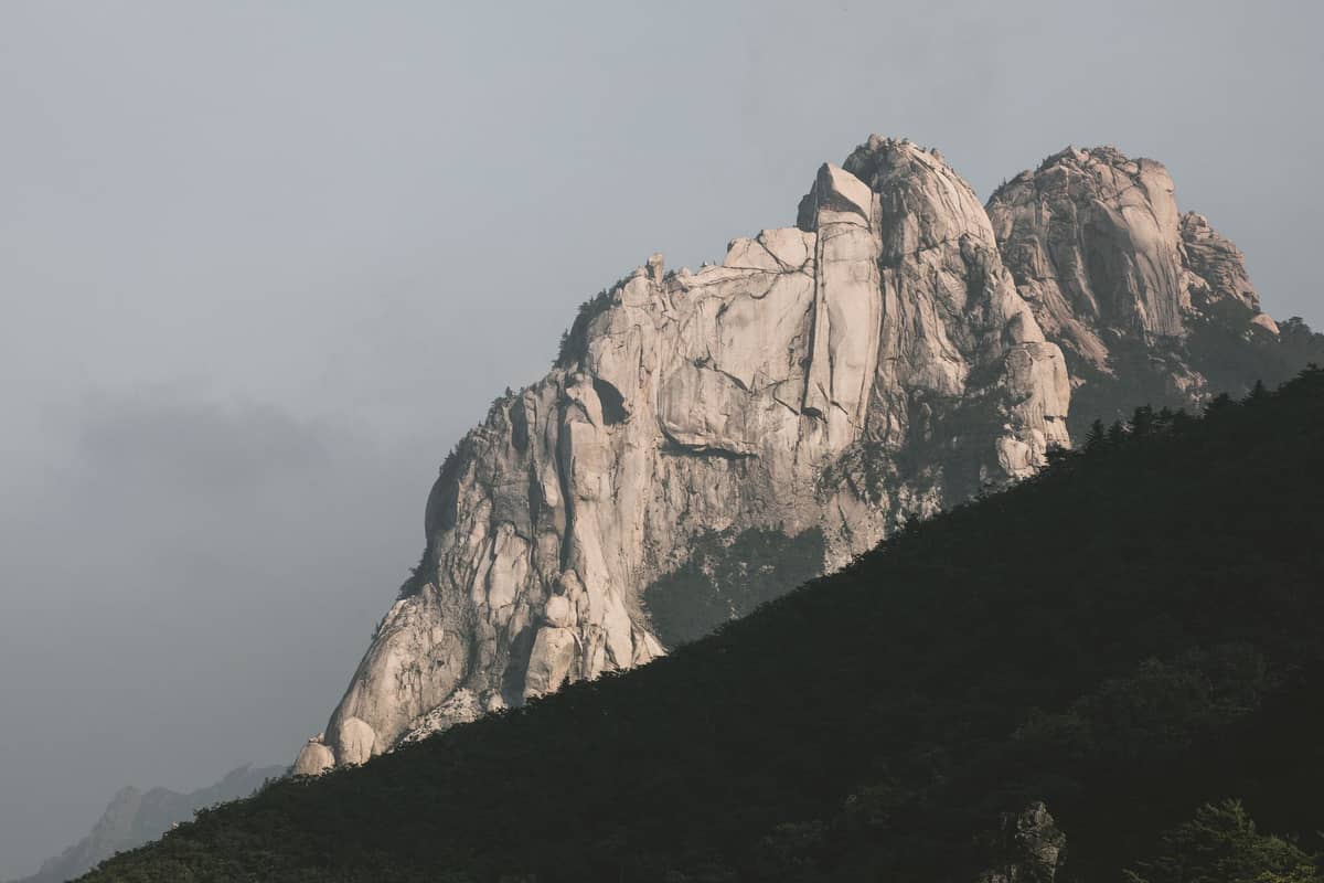 Picos de granito de uma montanha dramática erguendo-se acima de florestas verdes sob um céu nublado, característicos do Bukhansan em Seul.