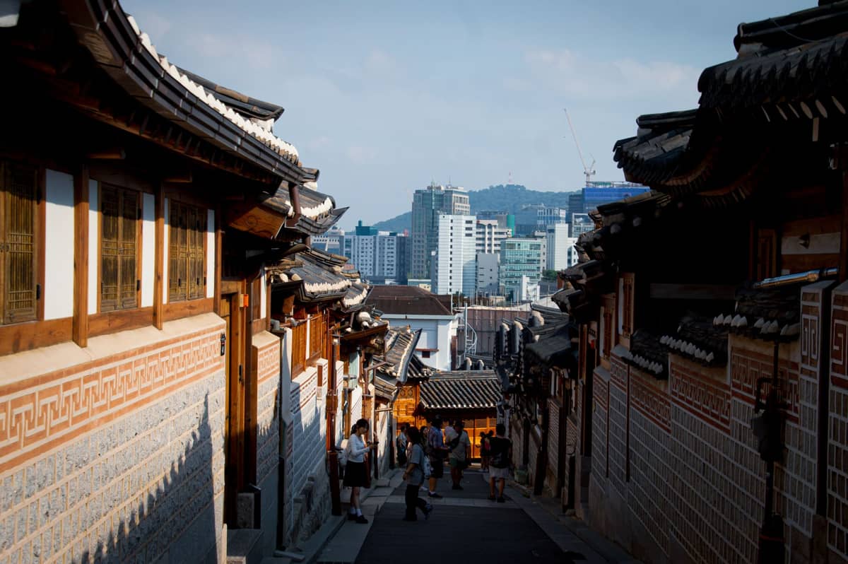 Rua íngreme e estreita no Bukchon Hanok Village de Seul com telhados hanok tradicionais, pedestres e edifícios da cidade visíveis ao fundo.