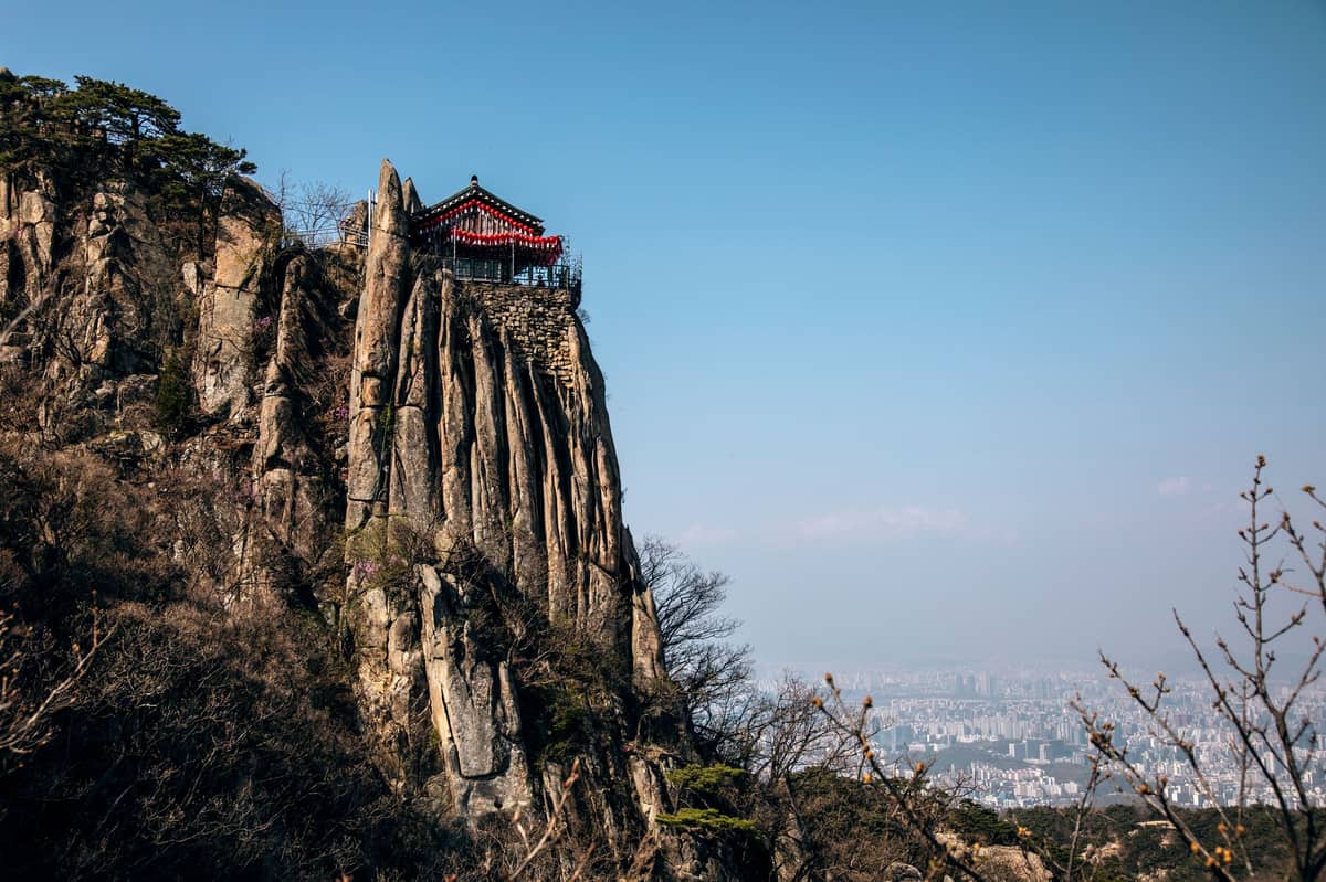 Um pavilhão coreano tradicional empoleirado sobre dramáticos penhascos rochosos com vista para o horizonte de Seul ao longe, combinando natureza, paisagens e patrimônio cultural.