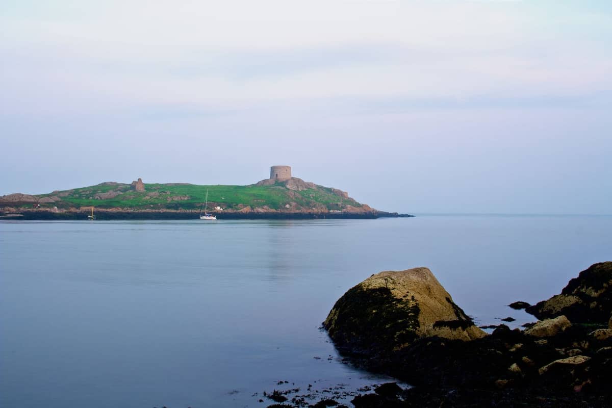Vue lointaine de Dalkey Island avec sa tour Martello, vue depuis le rivage avec une mer calme et des rochers au premier plan.