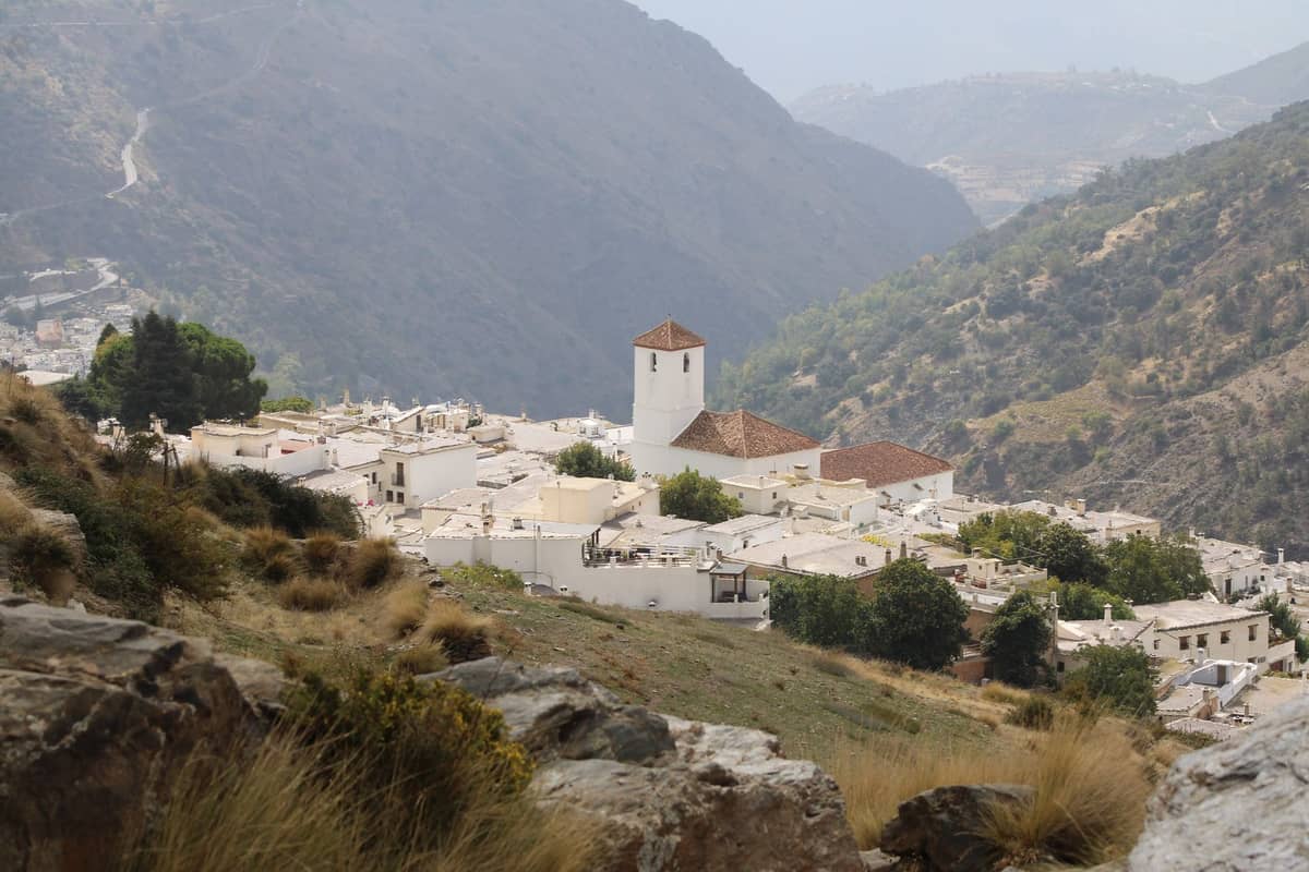 หมู่บ้านทาสีขาวพร้อมหอคอยโบสถ์ ตั้งอยู่บนเนินเขาลาดชันในแถบ Alpujarras ใกล้กับ Granada โดยมีเนินเขาขรุขระเป็นฉากหลัง