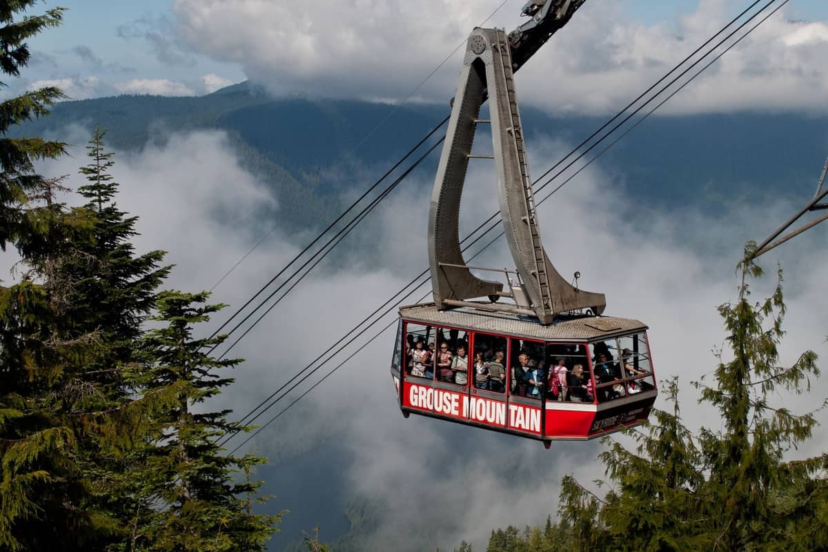 Eine Gondel der Grouse Mountain Aerial Tramway befördert Menschen hoch über Nadelbäume, mit den nebligen North Shore Mountains im Hintergrund.