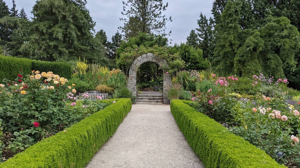 Weg durch einen gestalteten Garten mit üppigem Grün, blühenden Blumen und einem steinernen Torbogen, umgeben von hohen Bäumen.