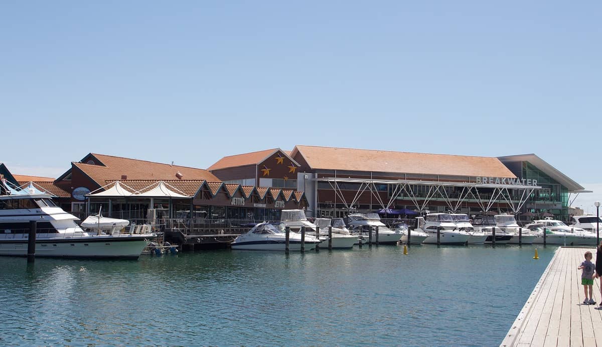 Blick auf den Hillarys Boat Harbour bei Tag: Boote liegen am Steg, im Hintergrund sind der Breakwater-Komplex, Restaurants und die Promenade zu sehen.