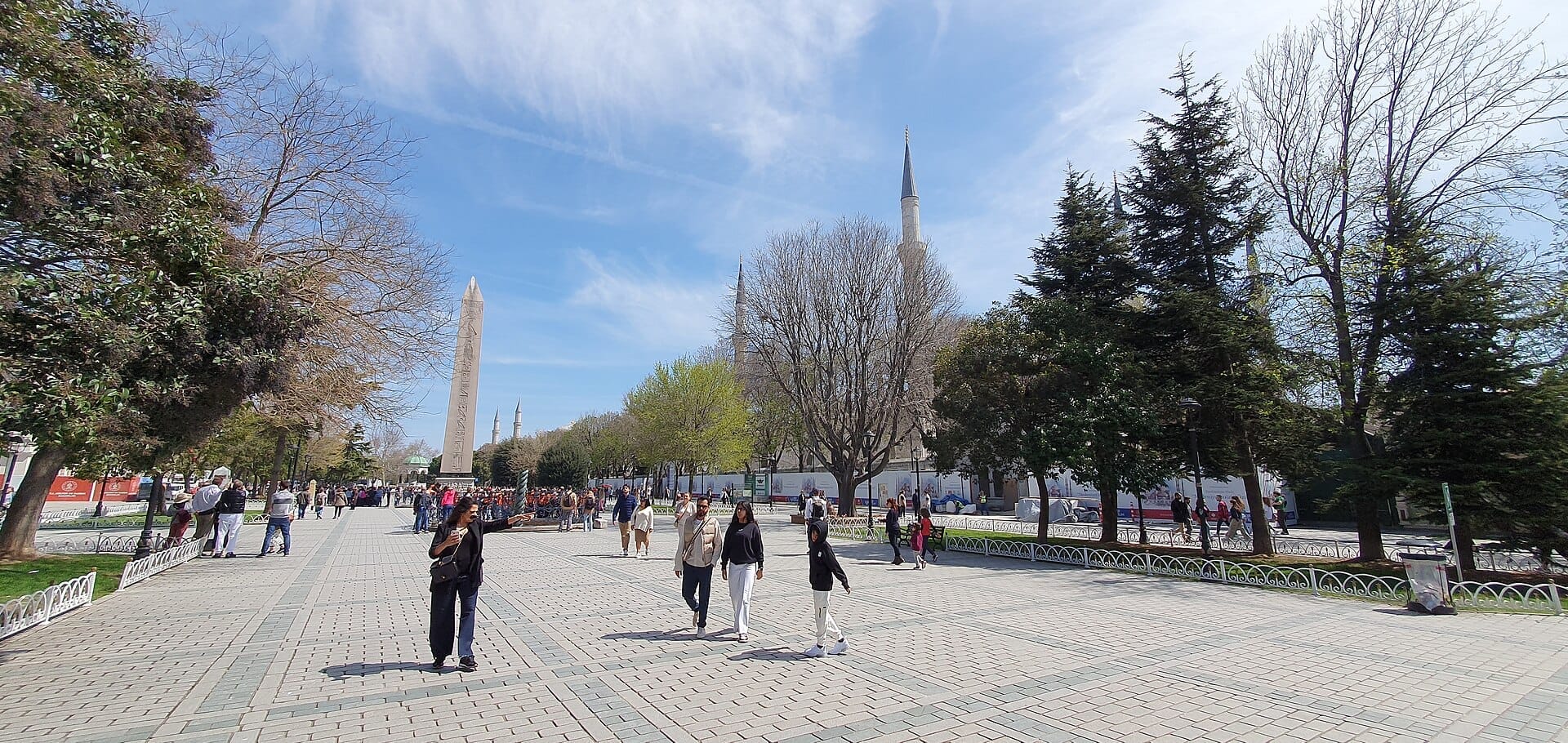 Ampia piazza in pietra all'Ippodromo di Costantinopoli, con l'antico Obelisco Egizio di Teodosio, minareti, alberi e persone che passeggiano sotto un cielo azzurro.