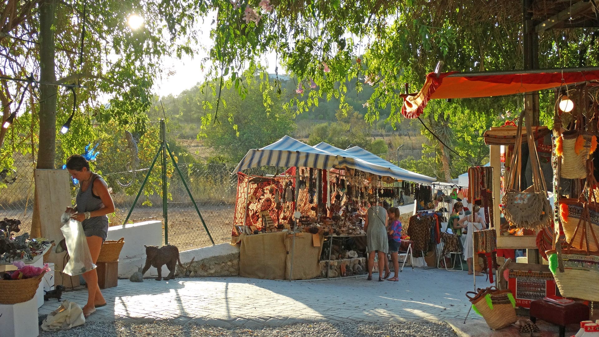Bancarelle all'aperto al Las Dalias Hippy Market di Ibiza espongono coloratissime borse artigianali, gioielli e oggetti di artigianato sotto tendoni a righe nel sole del pomeriggio.