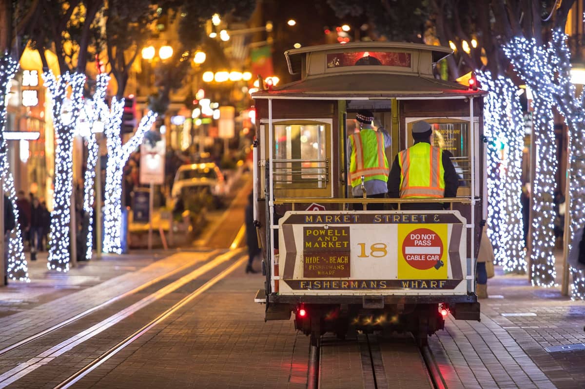 Teleférico de San Francisco con señalización de Powell & Market y Fisherman's Wharf, operado por trabajadores con chalecos de seguridad, iluminado por luces festivas en una calle de la ciudad de noche.