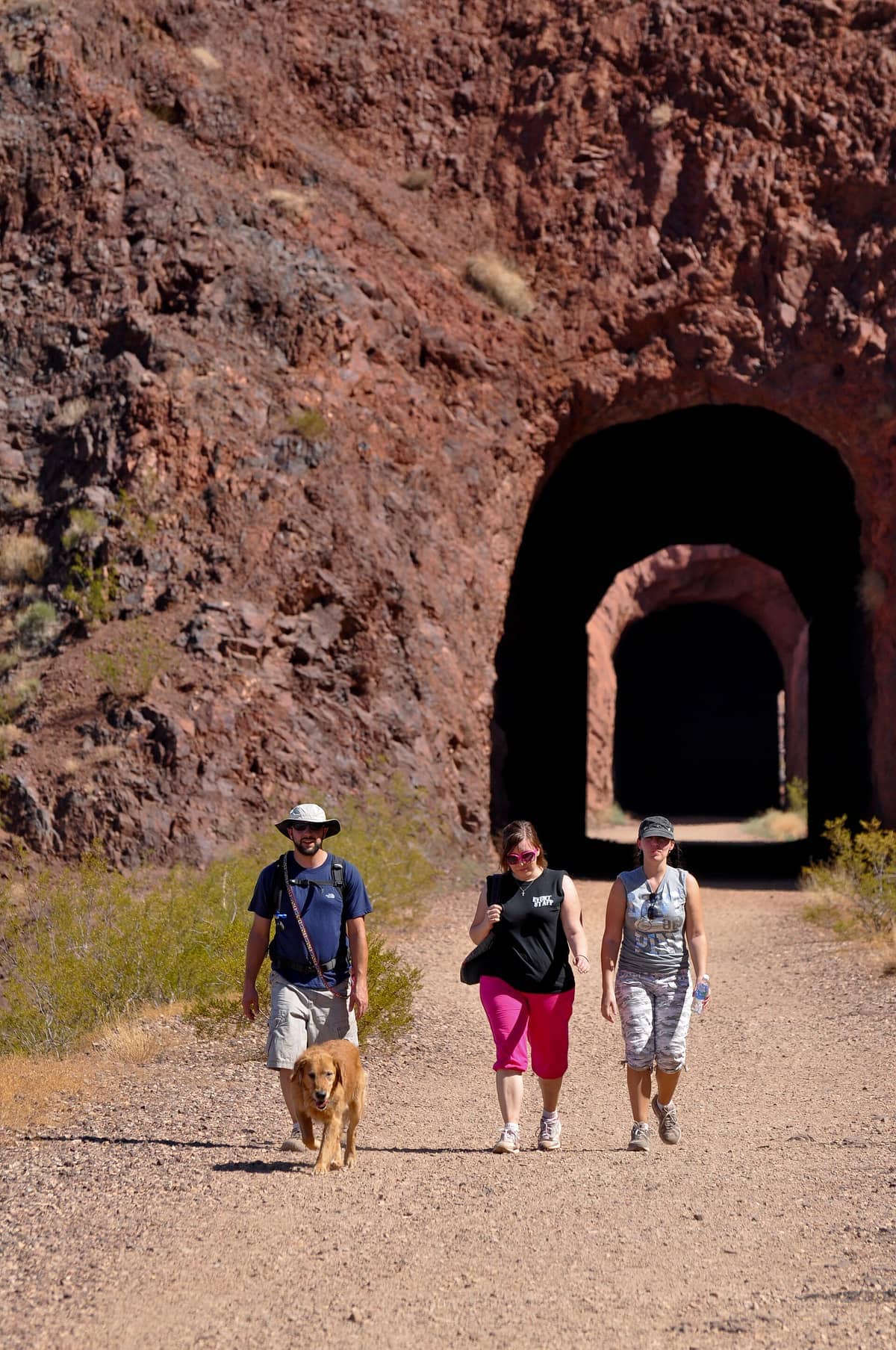 Tiga pendaki dan seekor anjing berjalan di sepanjang jalur kerikil melalui terowongan yang dibor dengan tangan di Historic Railroad Trail dekat Hoover Dam.