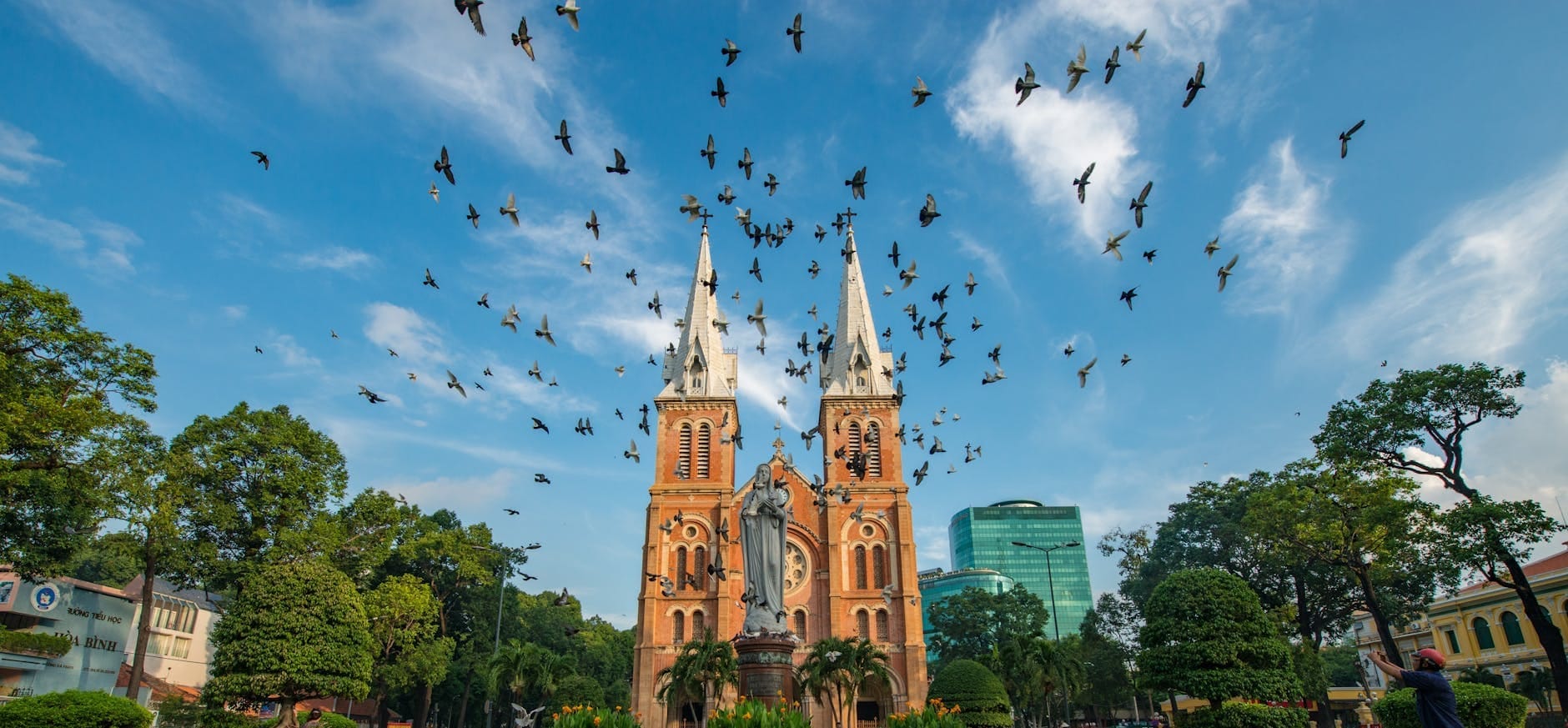 Catedral Basílica de Notre-Dame de Saigón con pájaros volando por encima, estatua de la Virgen María al frente, rodeada de vegetación y edificios de la ciudad.