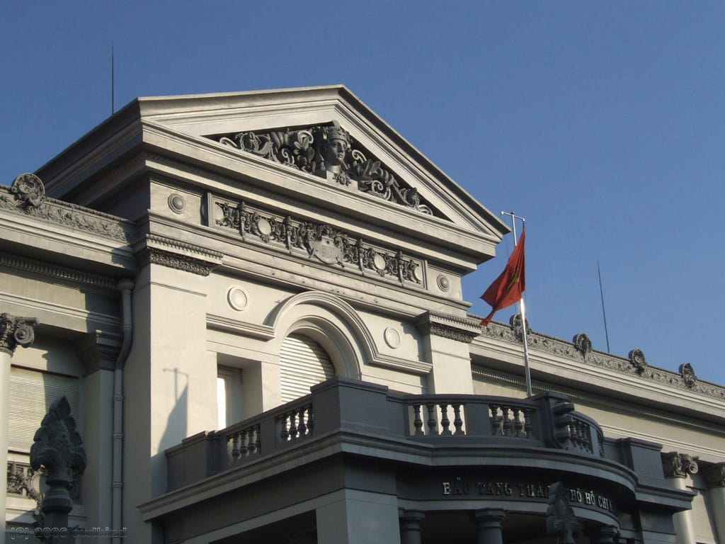 Fachada frontal del Museo de Ho Chi Minh con arquitectura colonial francesa, intrincadas tallas en piedra y una bandera vietnamita bajo un cielo azul despejado.