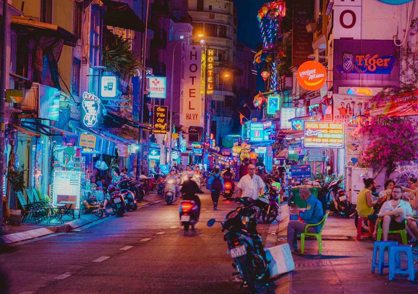 Concurrida calle Bùi Viện de noche con luces de neón, bares, comida callejera y personas sentadas en taburetes azules en la acera.
