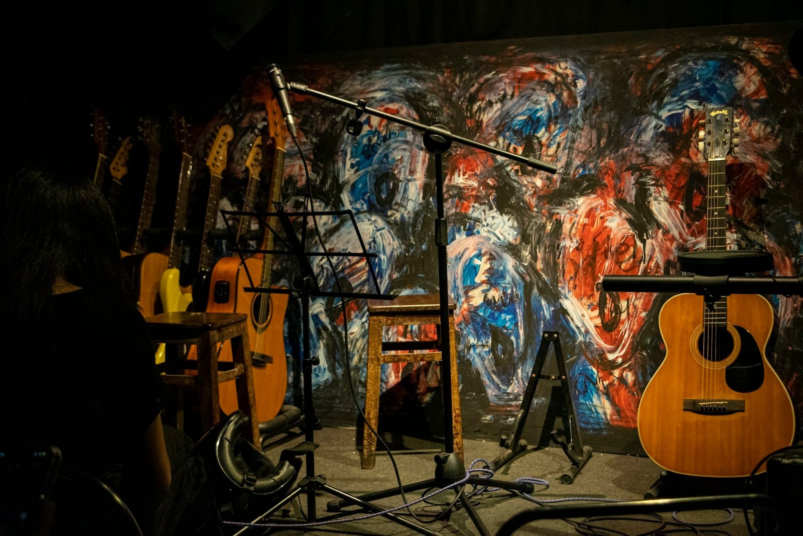 Escenario íntimo de música en vivo con múltiples guitarras, soportes para micrófono, taburetes y colorido mural abstracto de fondo, listo para una actuación.