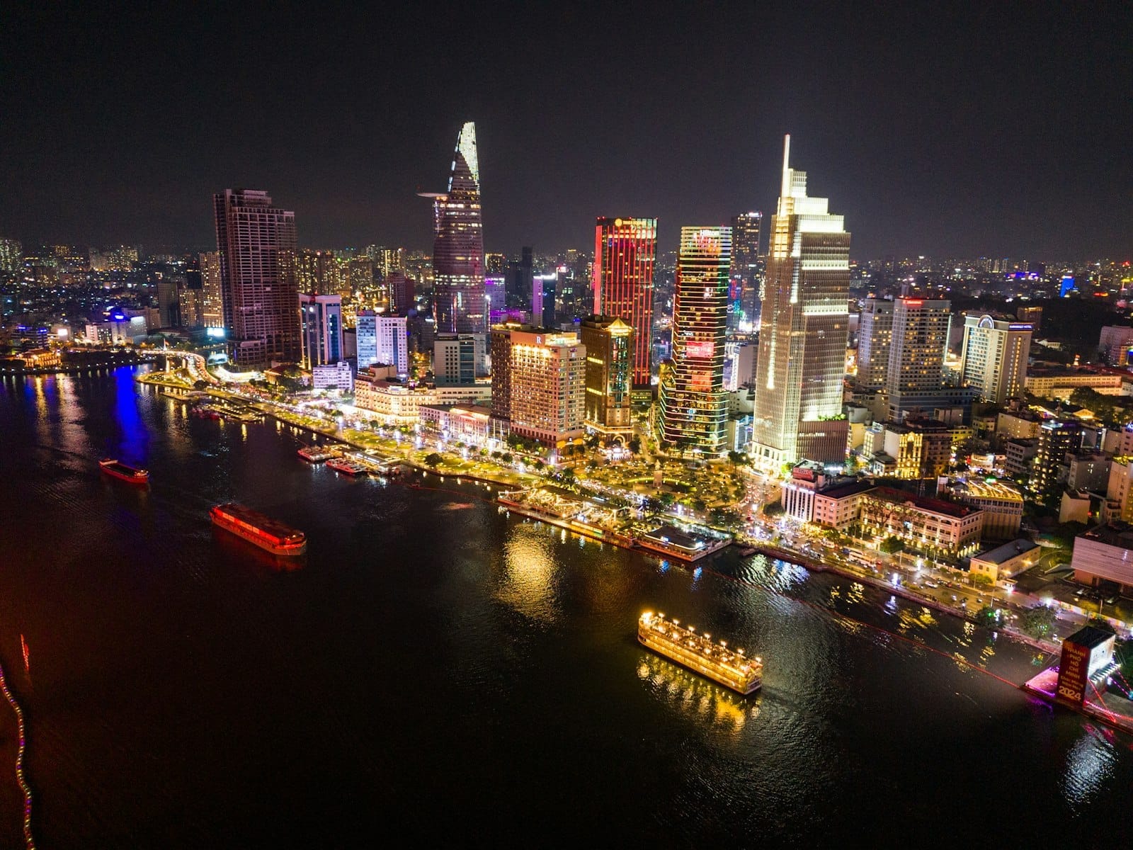 Vista aérea vibrante del malecón iluminado de Ho Chi Minh City de noche, con rascacielos de colores, barcas animadas y una intensa vida nocturna junto al agua.