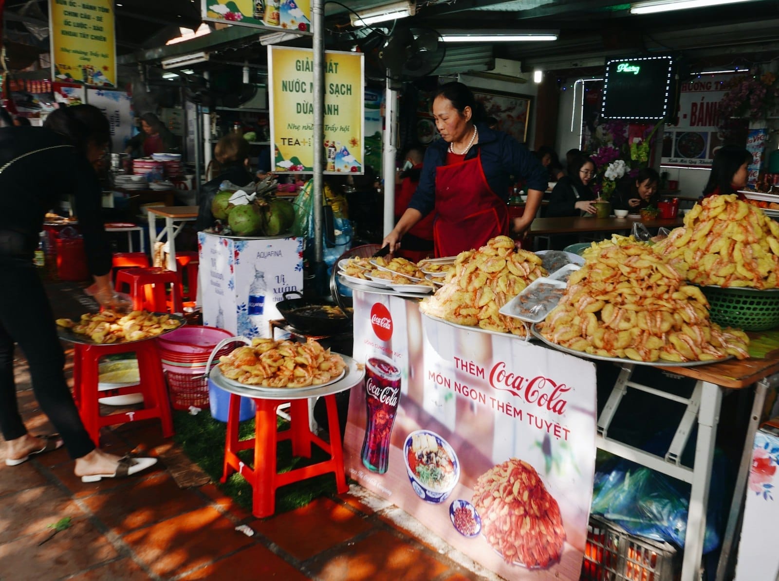 Vendedora de comida callejera en Ho Chi Minh City frente a mesas repletas de tortillas vietnamitas saladas, rodeada de carteles coloridos y taburetes pequeños.