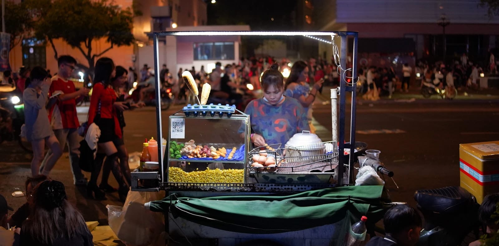 Vendedor de comida callejera asando brochetas en un animado mercado nocturno en Ho Chi Minh City, rodeado de personas y brillantes luces de la ciudad.