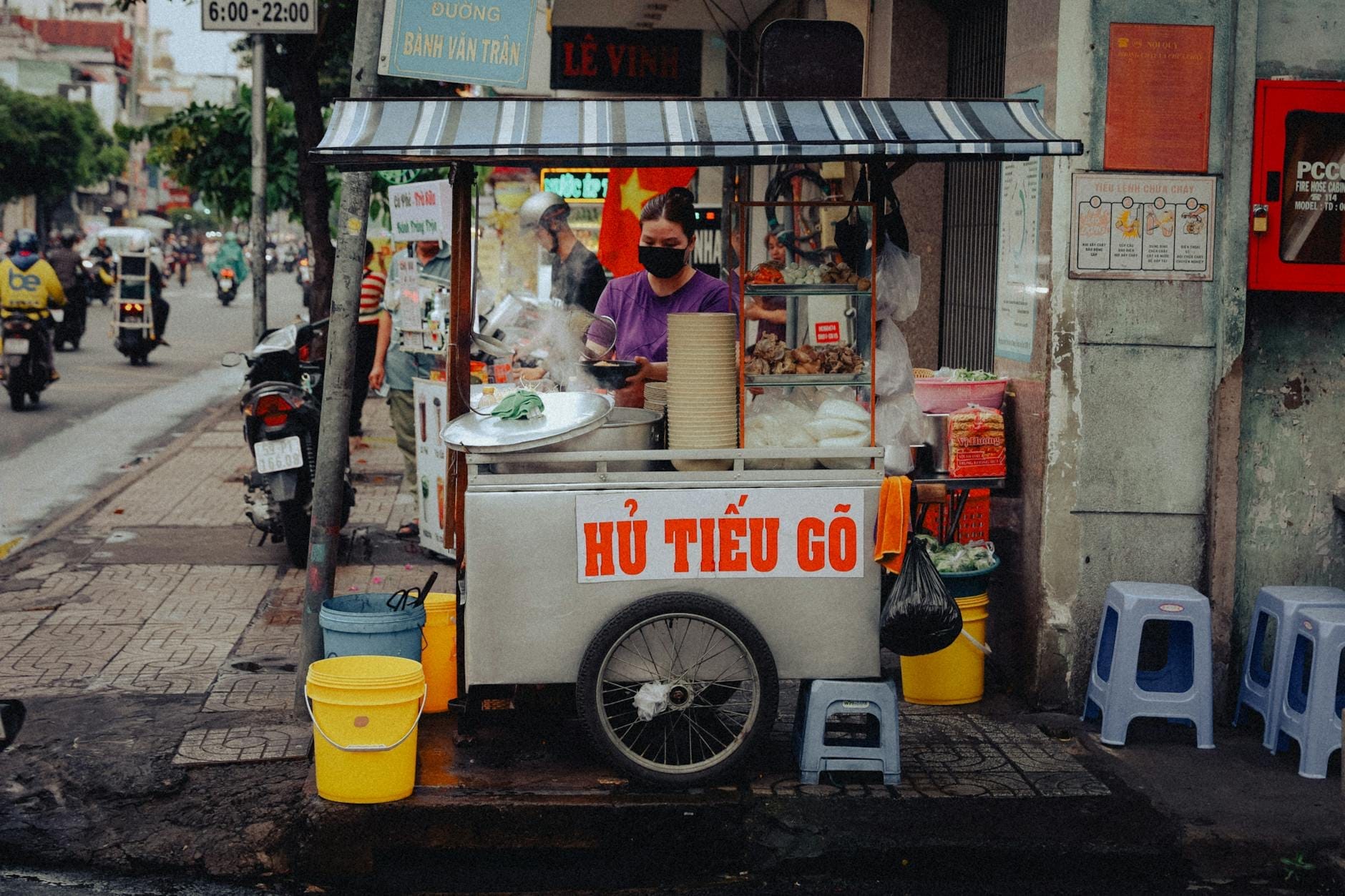 Un vendedor ambulante prepara fideos en un carrito en la acera de Ho Chi Minh City, con letreros en vietnamita y paisaje típico de la calle local.