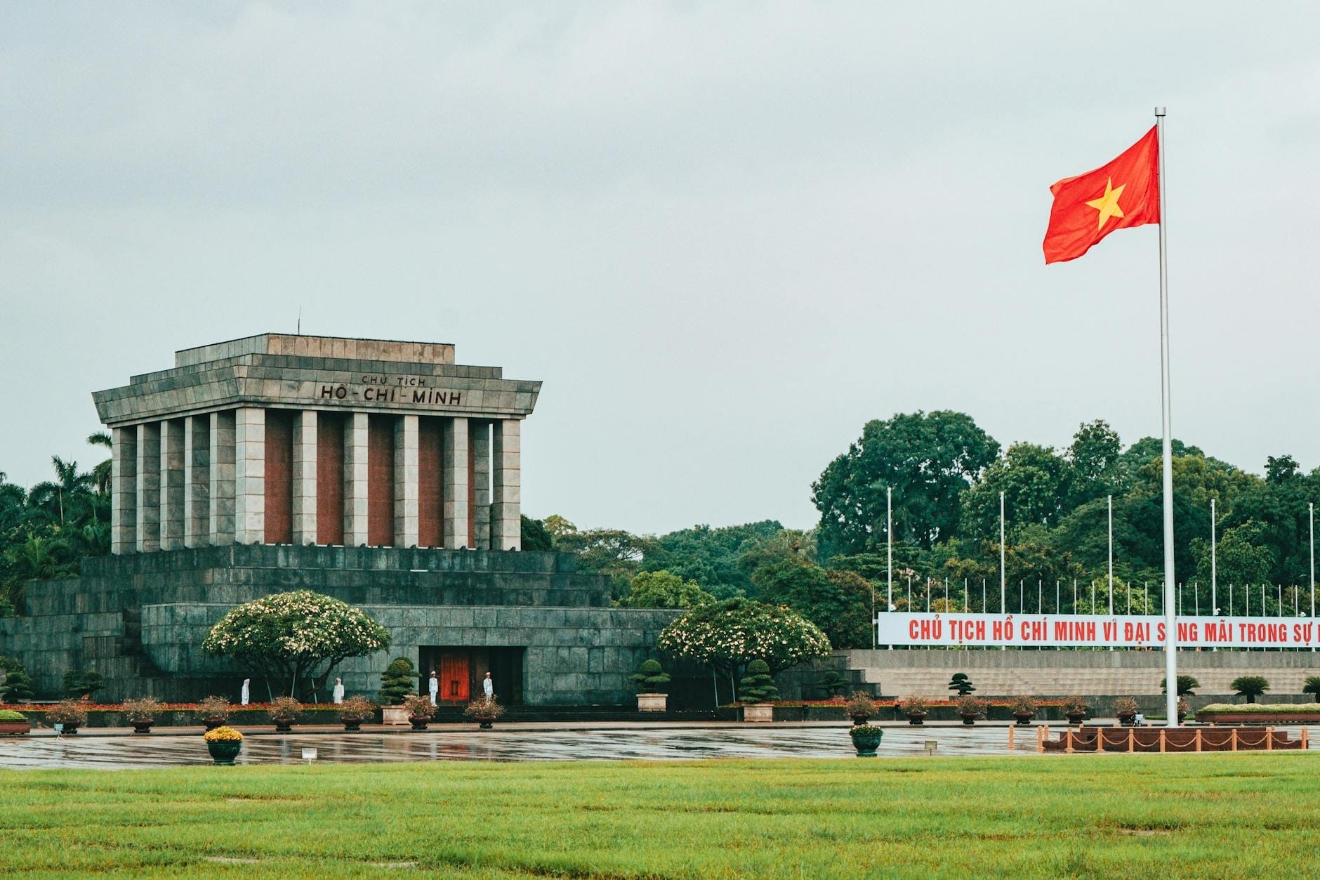 Wide landscape view of Ho Chi Minh Mausoleum in Hanoi with Vietnamese flag and surrounding gardens on a cloudy day.