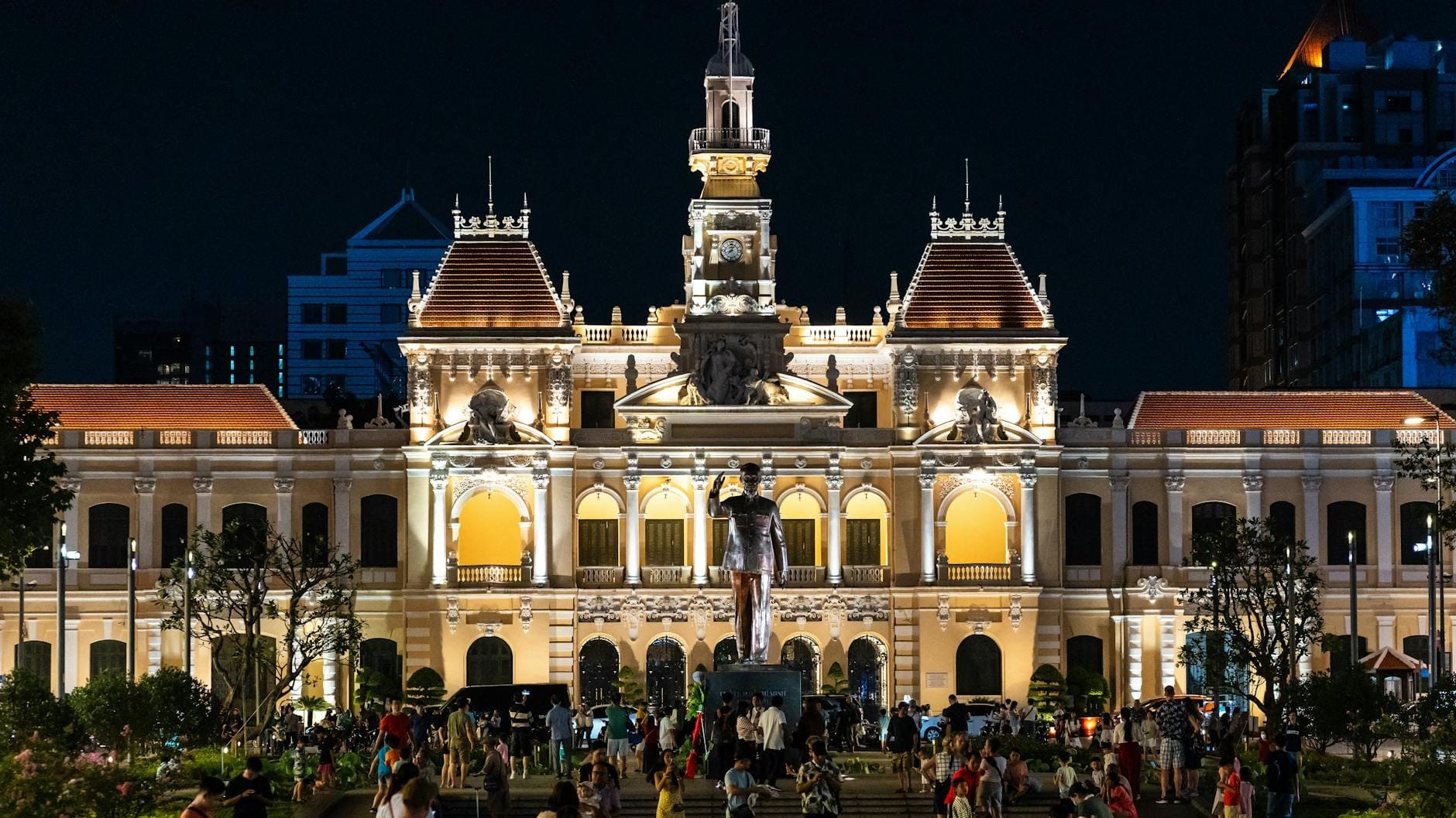 Vista nocturna de la estatua de Ho Chi Minh frente al iluminado Edificio del Comité Popular en el Paseo Peatonal Nguyen Hue, con un ambiente muy animado y mucha gente disfrutando de la noche.