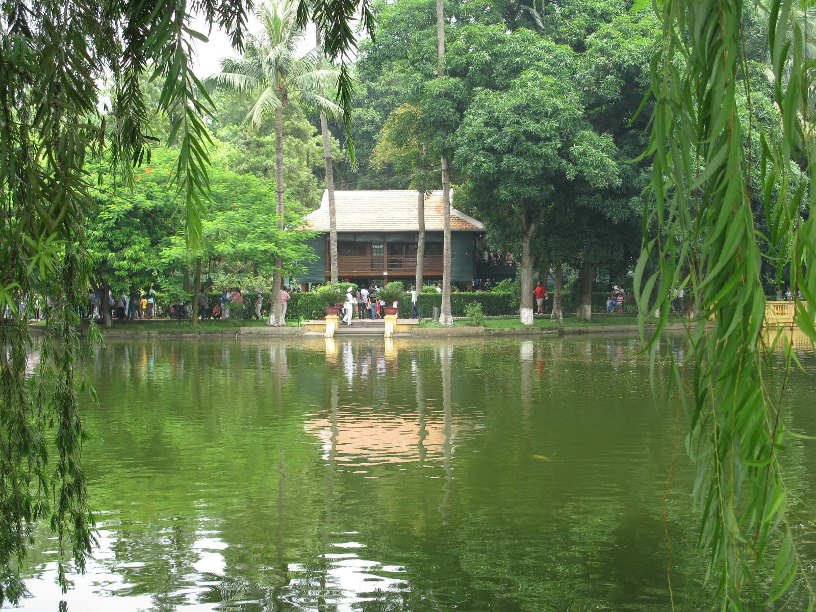 Ho Chi Minh's Stilt House, a simple wooden building, stands beside a peaceful pond surrounded by lush green trees in Hanoi.