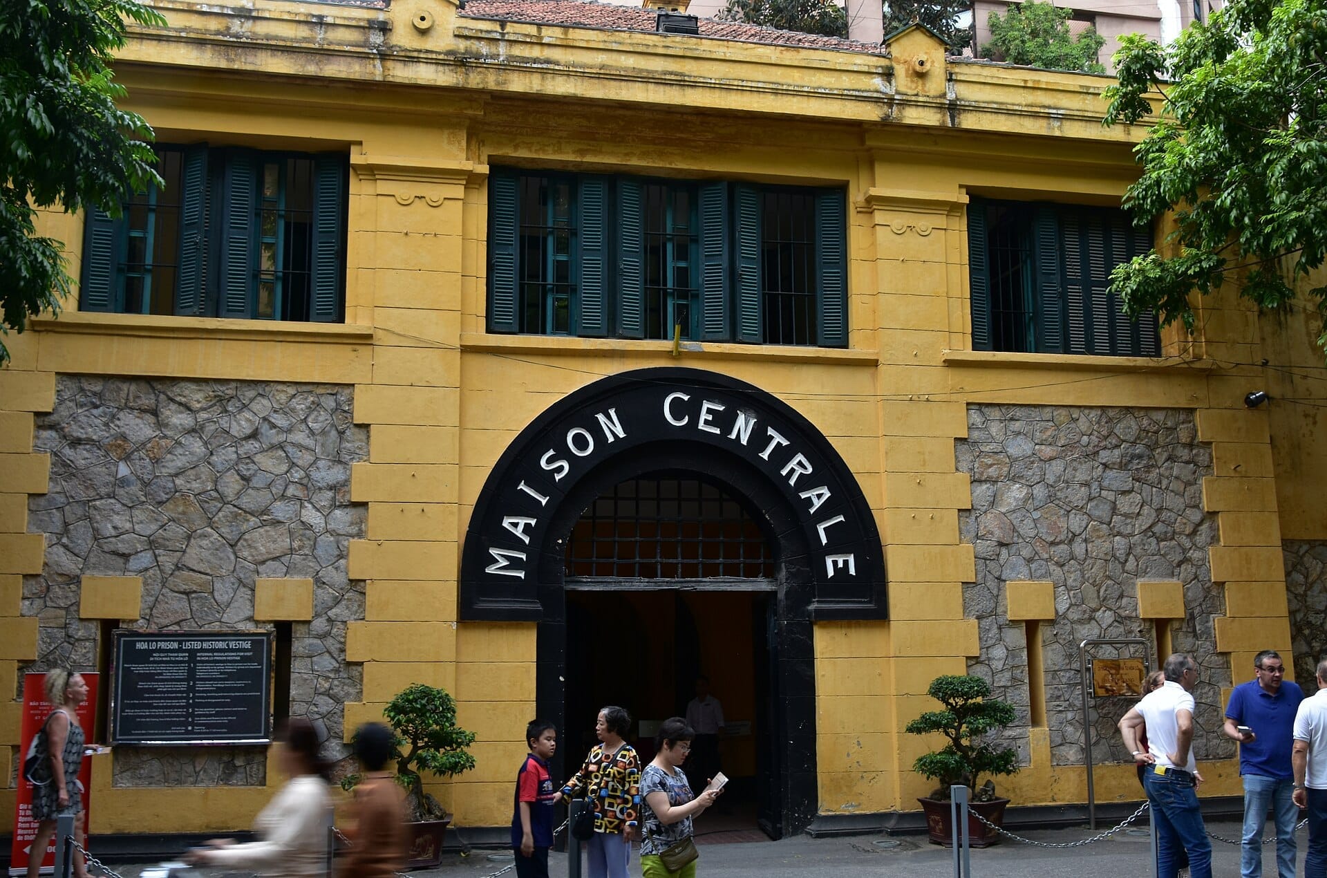 Front entrance of Hoa Lo Prison in Hanoi, featuring yellow walls, barred windows, and visitors mingling outside under green trees.