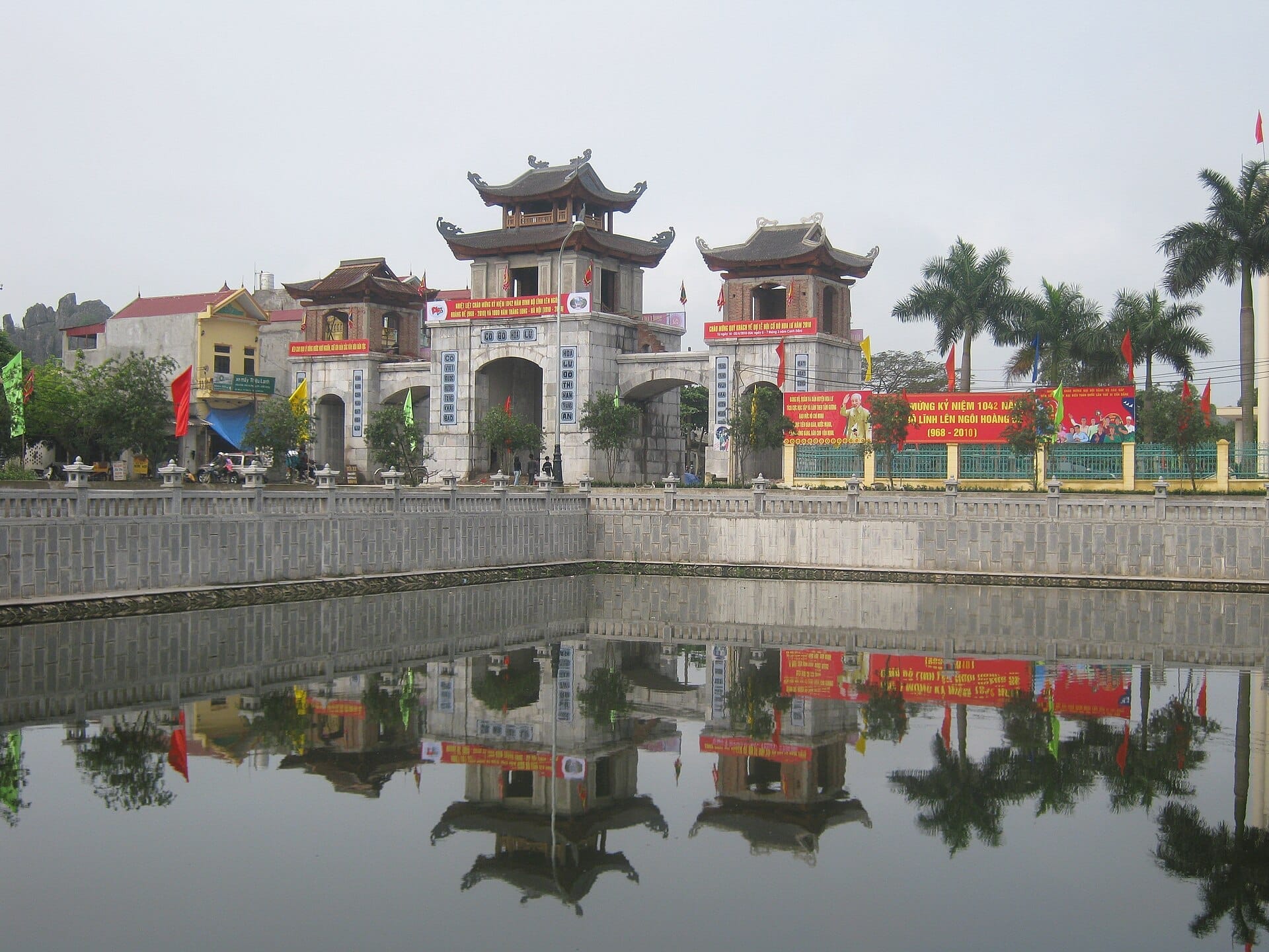 Main entrance gate of Hoa Lư Ancient Capital reflected in a tranquil pond, surrounded by palm trees, colorful banners, and traditional Vietnamese architecture.