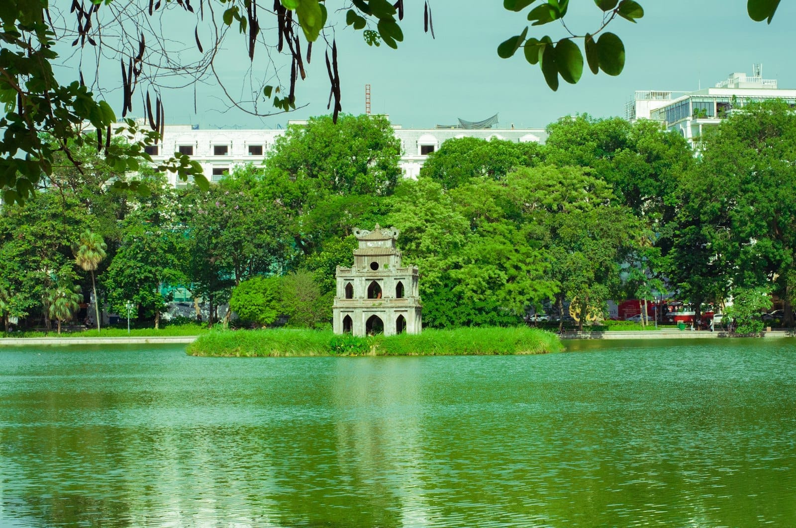 View of Hoàn Kiếm Lake with Turtle Tower on the small island, surrounded by trees and city buildings in the background, framed by leafy branches.