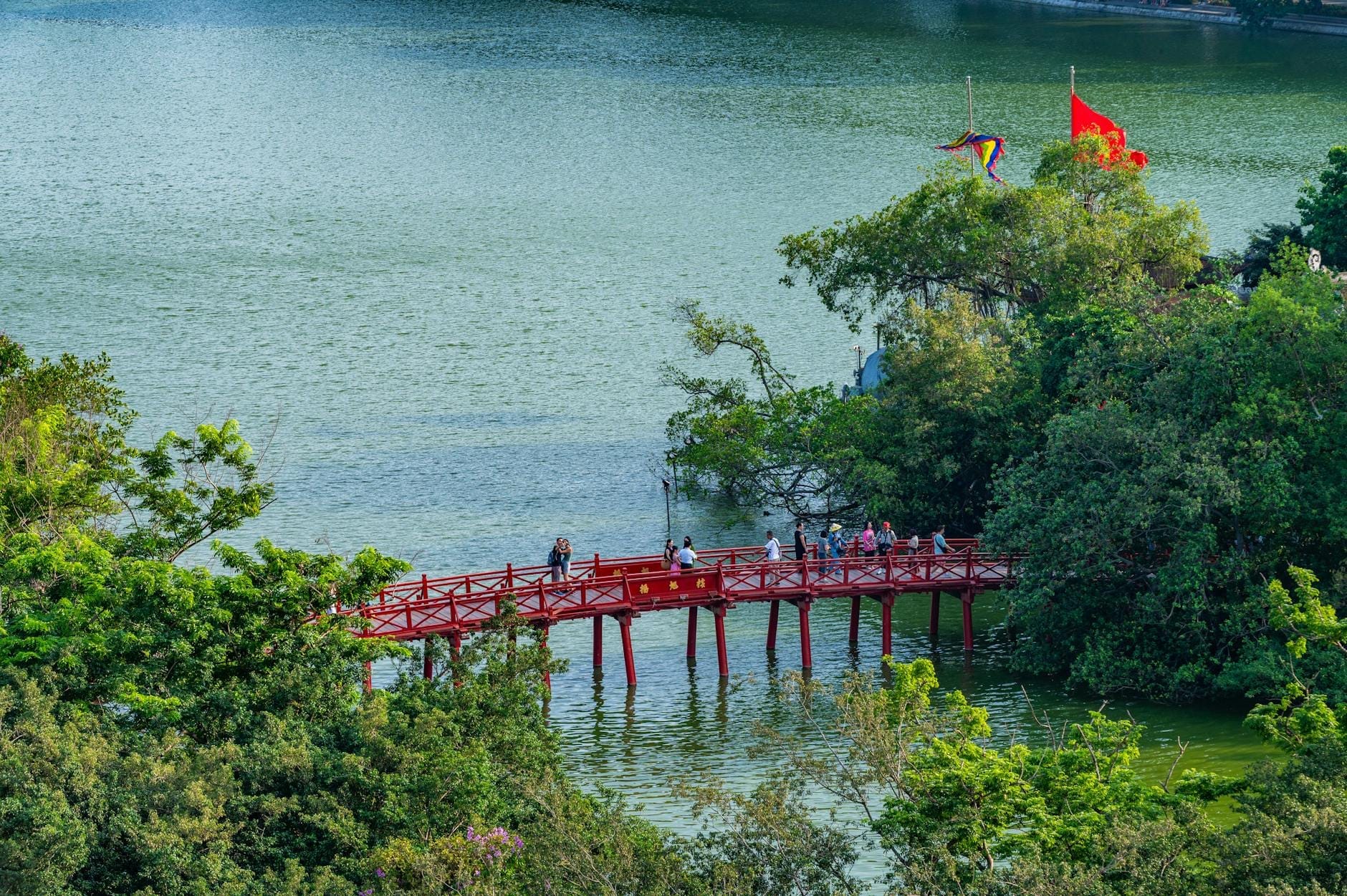 An aerial view of Hoàn Kiếm Lake with the iconic red-painted Huc Bridge stretching across the water toward a tree-covered temple, framed by vibrant greenery and Vietnamese flags.