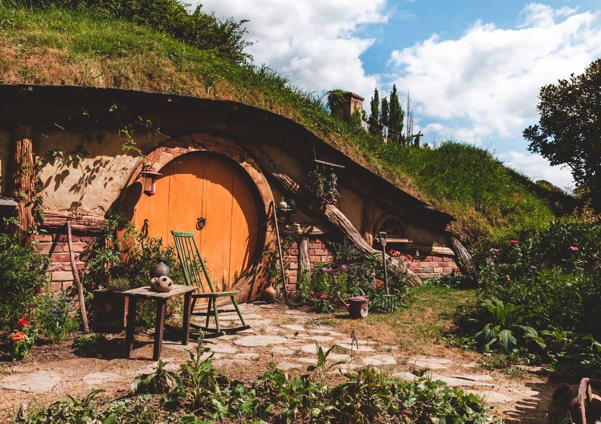 Ein malerisches Hobbit-Loch mit einer runden orangefarbenen Tür in einem grünen Hügel, umgeben von Gärten und rustikalen Gartenmöbeln.