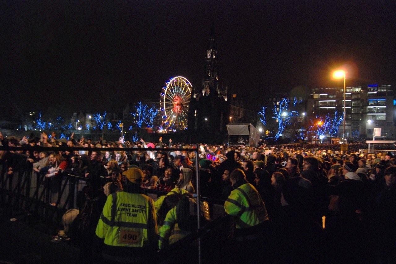Große Menschenmassen bei Nacht auf der Hogmanay-Straßenparty in Edinburgh, mit einem beleuchteten Riesenrad, blauen Festlichtern und dem Scott Monument im Hintergrund.