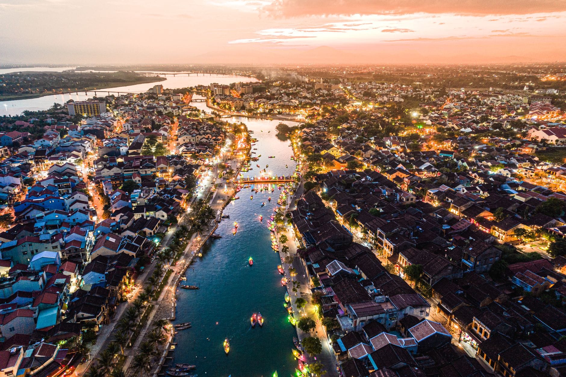 วิวทางอากาศยามพระอาทิตย์ตกของ Hoi An Ancient Town และแม่น้ำที่เรียงรายด้วยเรือประดับโคมไฟและอาคารริมน้ำที่เรืองแสง