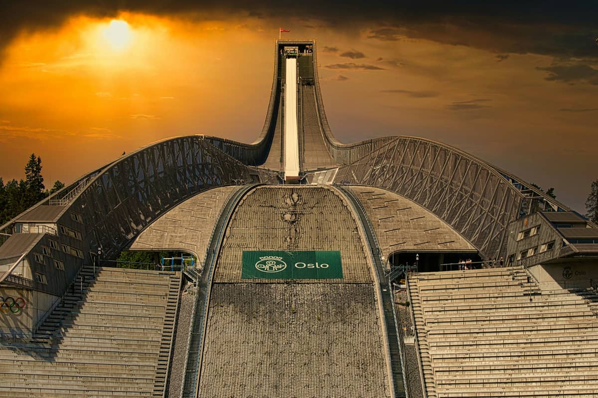 ภาพโคลสอัพจุดสกีจัมป์ชื่อดัง Holmenkollen ในออสโลกับท้องฟ้ายามพระอาทิตย์ตก เห็นทางลาดสกีและสัญลักษณ์ออสโลในบรรยากาศฤดูร้อน