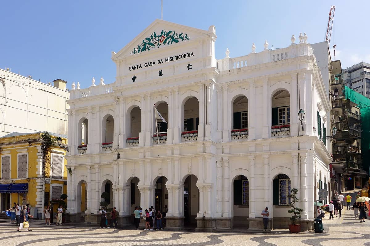 Das Heilige Haus der Barmherzigkeit in Macau: ein weißes neoklassizistisches Gebäude mit Bogenfenstern, portugiesischen und chinesischen Schriftzügen, davor der gemusterte Senado-Platz mit Passanten.