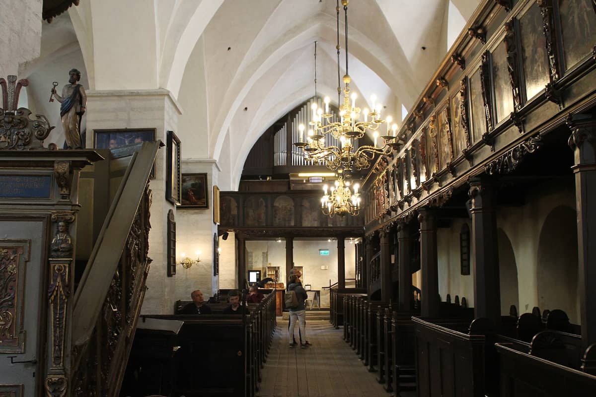 Innenansicht der Heilig-Geist-Kirche in Tallinn mit Holzbänken, prunkvollen Kronleuchtern, historisch bemalter Empore und gotischen Gewölbedecken.