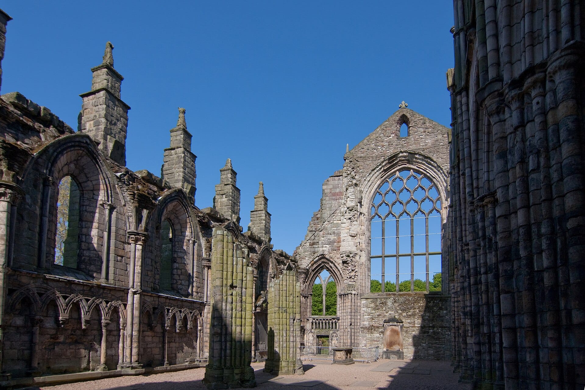 Innenansicht der Holyrood Abbey Ruinen mit aufragenden Gotikbögen und großen Fensteröffnungen, durch die ein strahlend blauer Himmel zu sehen ist.