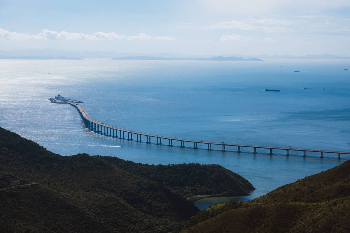 Weitblick auf die Hongkong–Zhuhai–Macao-Brücke, die sich über blaues Wasser in Richtung einer künstlichen Insel erstreckt, umgeben von Hügeln unter klarem Himmel.