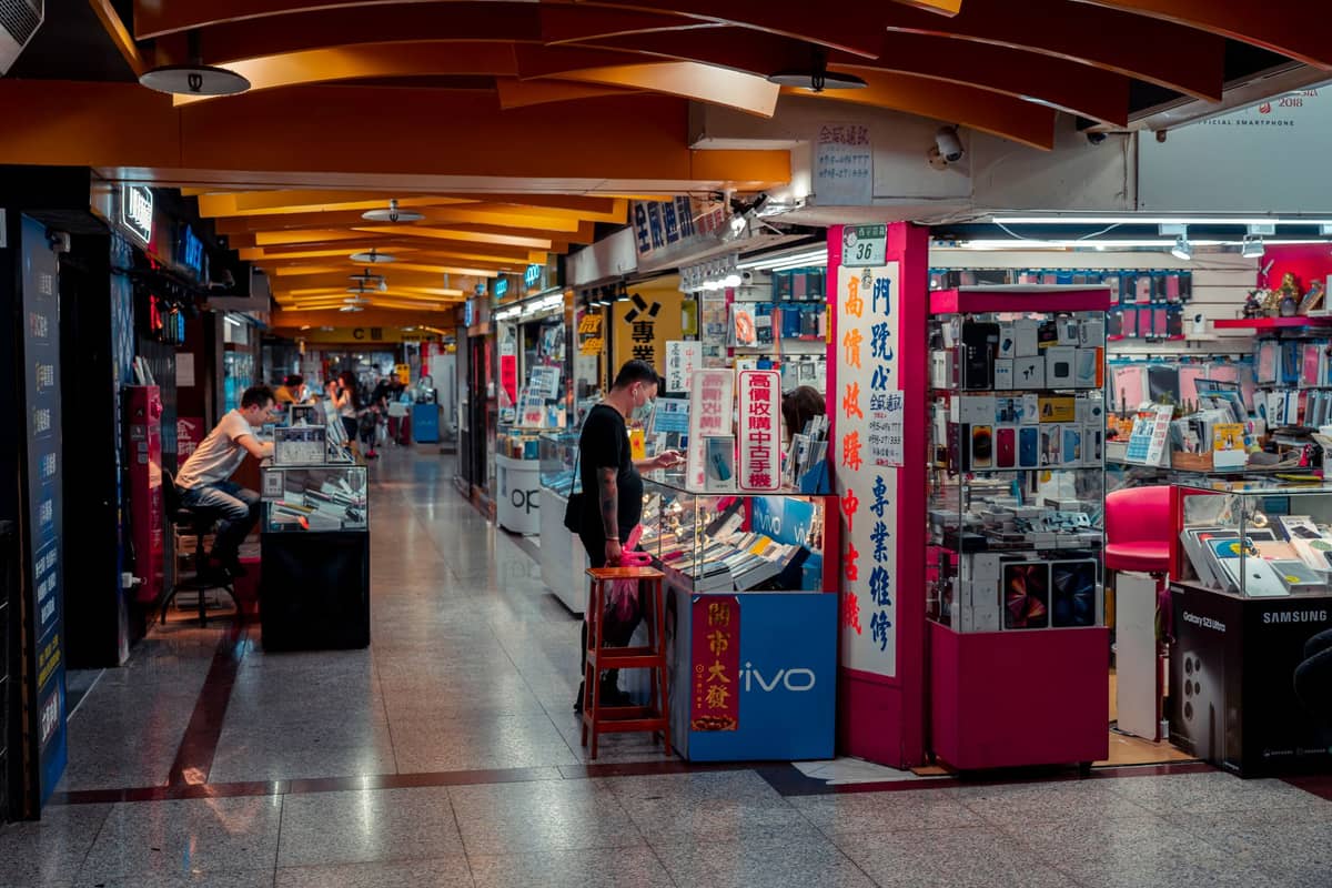 Interior de um mercado de eletrônicos na China, com vários estandes de acessórios para celulares, exposições de mercadorias e pessoas explorando os itens.