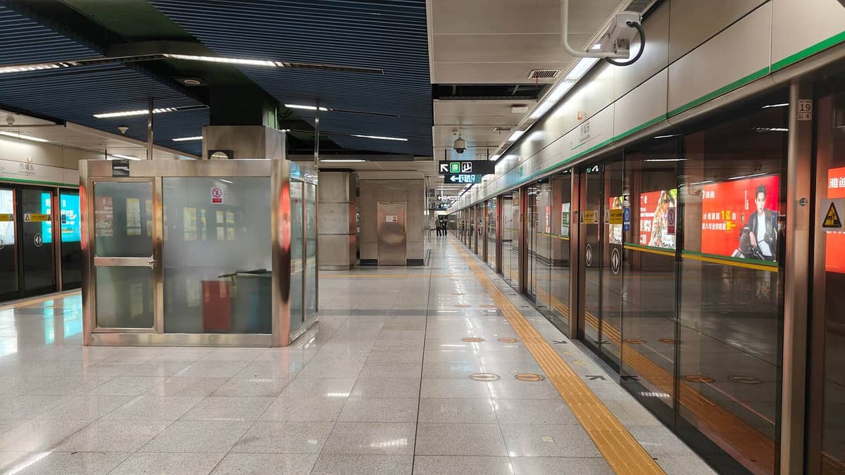 Vista ampla da plataforma de uma estação moderna do Metrô de Shenzhen com portas de vidro, sinalização e anúncios iluminados, mostrando o ambiente típico da estação para os viajantes.