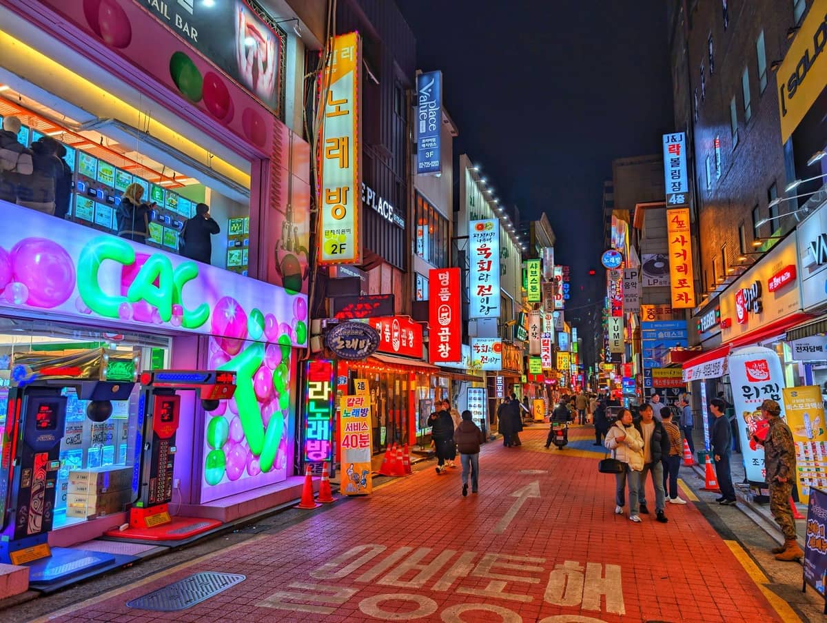 Cena noturna em Hongdae e Mapo com letreiros de néon iluminados, fachadas coloridas e pessoas passeando por uma animada área de pedestres.