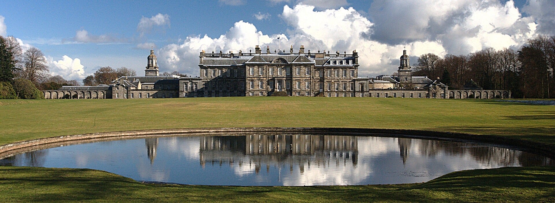 Weitwinkelpanorama von Hopetoun House mit seiner prachtvollen Barockfassade, die sich in einem Zierbecken spiegelt, satte Rasenflächen und Bäume unter einem teils bewölkten Himmel.