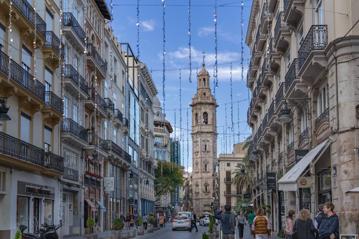 Una animada calle de Valencia con personas caminando, flanqueada por cafés y tiendas, que conduce a una histórica torre campanario bajo un cielo azul.