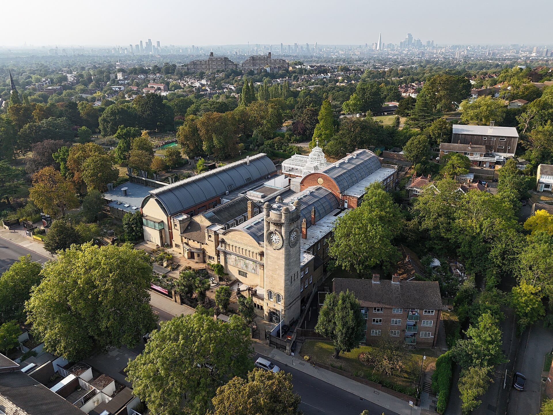 Vista aerea dell'Horniman Museum and Gardens circondato da una rigogliosa vegetazione, con lo skyline del centro di Londra visibile nella nebbiosa distanza.