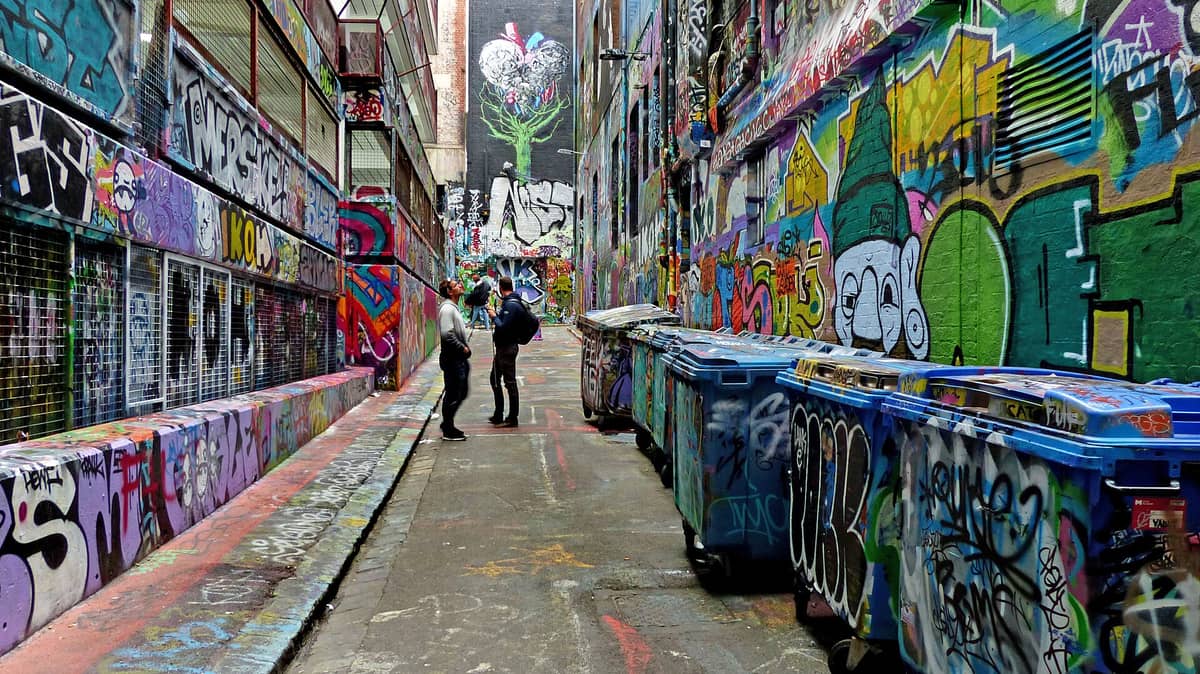 Un callejón amplio y colorido con arte urbano y grafitis vibrantes en ambas paredes, donde varias personas admiran los murales y grandes contenedores de basura se alinean a un lado. Luz de día, atmósfera urbana.