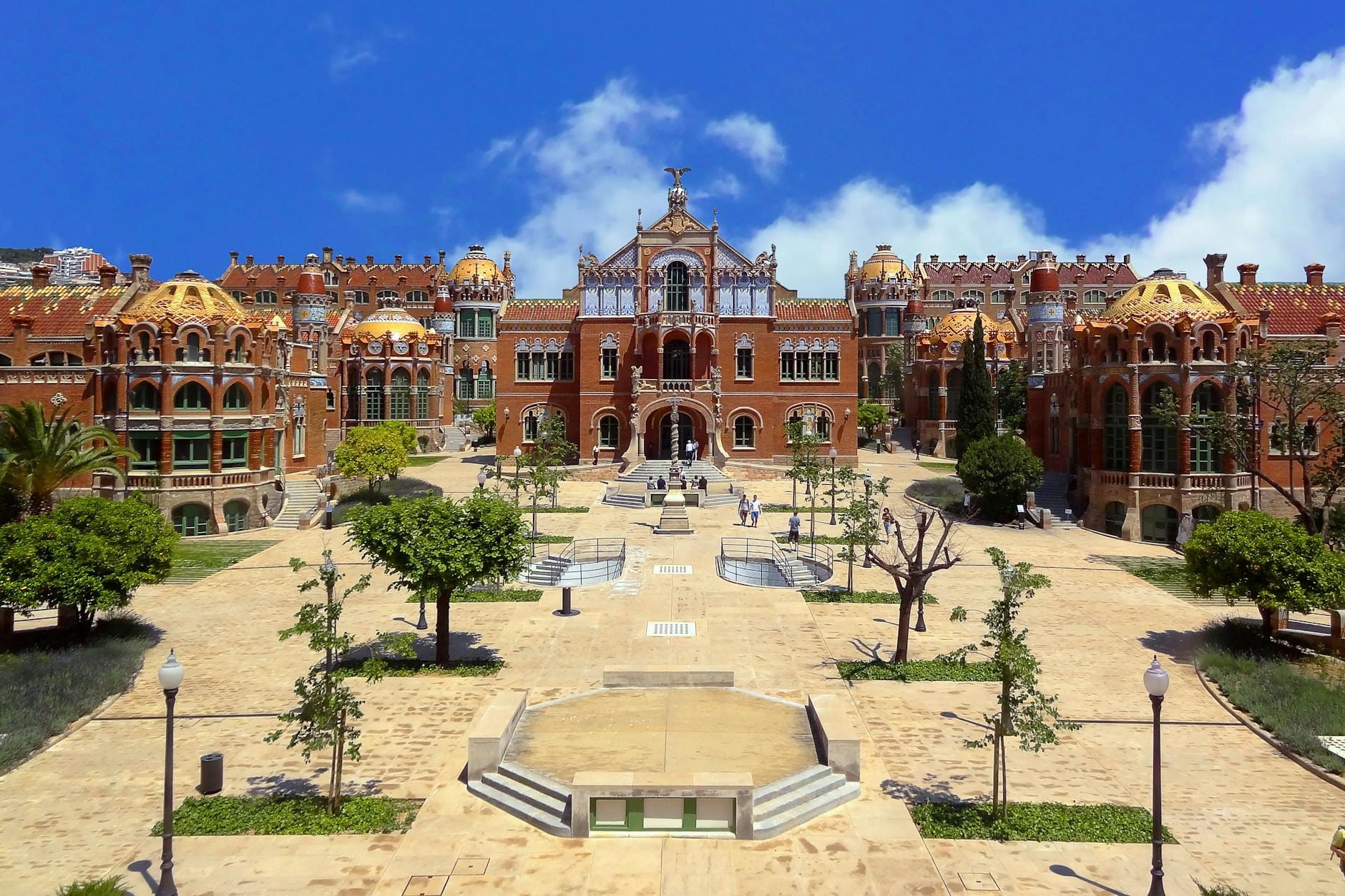 Wide view of the Hospital de Sant Pau complex in Barcelona under a bright blue sky, showcasing its modernista architecture, domes, and central courtyard.