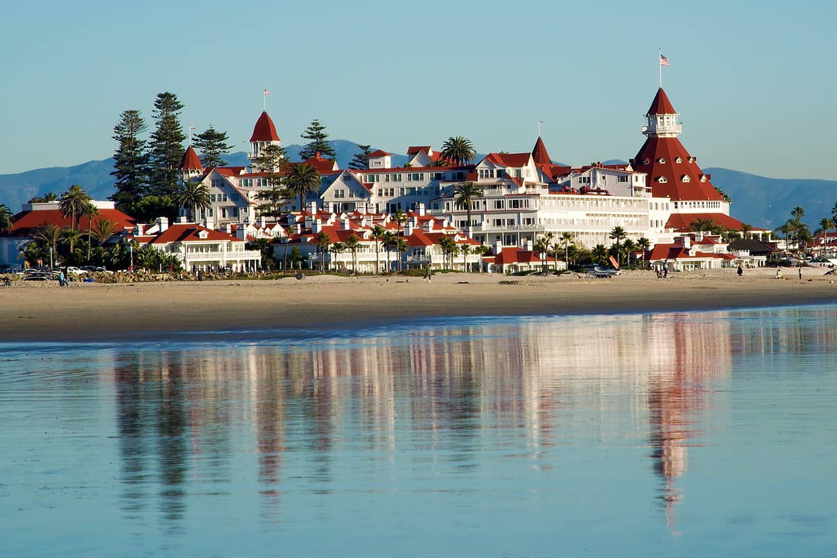 วิวอาคารสีแดงทรงวิคตอเรียอันเป็นเอกลักษณ์ของ Hotel del Coronado มองจากชายหาดในวันแดดจ้า มีต้นปาล์มและเงาสะท้อนบนทรายเปียก