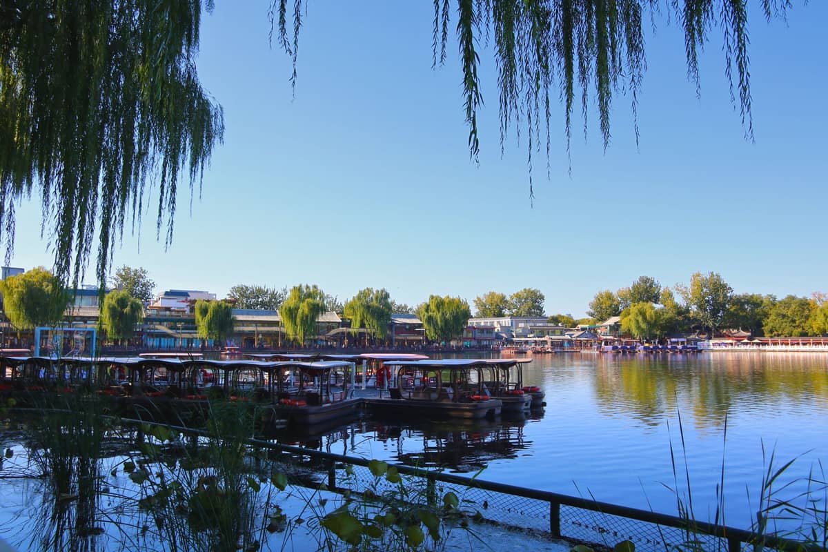Weiter Blick auf den Houhai oder Shichahai-See mit Weiden, vertäuten Booten und Ufergebäuden unter blauem Himmel in Peking.