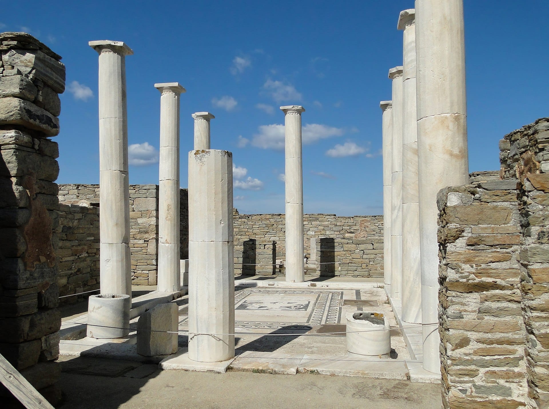Columnas de mármol y antiguas paredes de piedra rodean el famoso patio de mosaicos en la Casa de Dioniso en Delos bajo un cielo azul despejado.