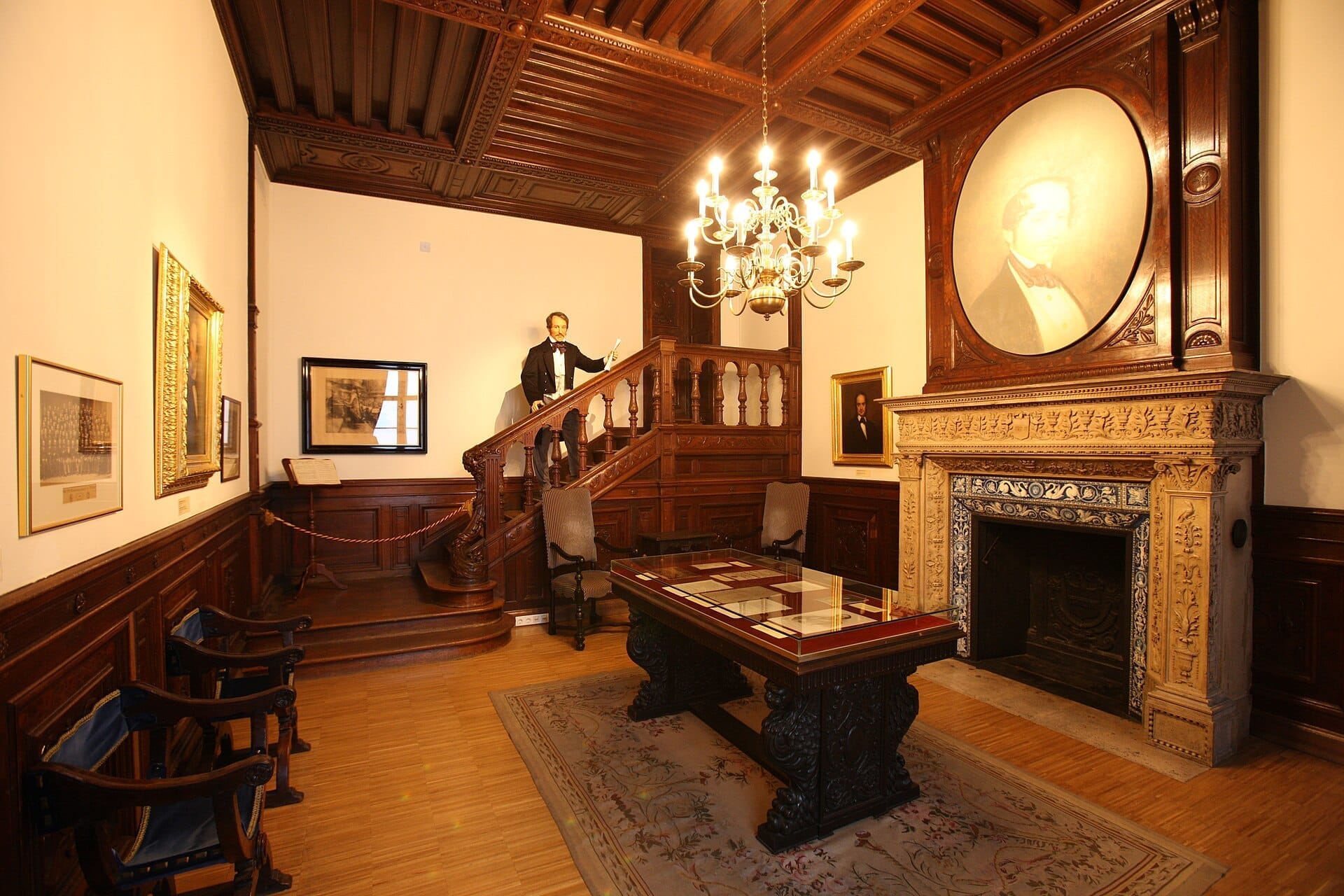 Elegantes holzgetäfeltes Zimmer im Haus der Musik in Wien mit einem Kronleuchter, Porträts, einem großen Kamin und einer Treppe mit einer lebensechten Figur.
